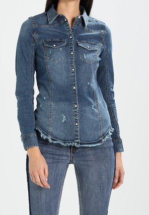 Vrouw draagt een aangesloten, geknoopt blauw denim overhemd met borstzakken en verweerde details, gecombineerd met tweekleurige blauwe jeans.