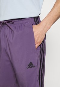 Fialové sportovní šortky vyrobené z lehkého materiálu, s černým pasem, černými bočními pruhy a vyšitým černým logem.