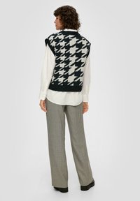 Gilet en tricot noir et blanc à motif pied-de-poule, sans manches, porté sur une chemise blanche ; associé à un pantalon à carreaux taille haute.