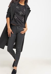 Top negro bordado con acentos plateados, debajo de un abrigo largo negro, combinado con pantalones ajustados negros y zapatos negros.