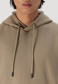 Tan hoodie with a hood, featuring long drawstrings with black tips. Soft, textured fabric, designed for comfort and casual wear.