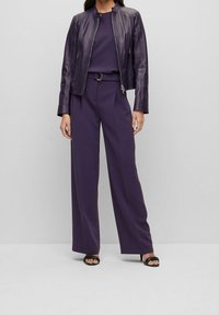 Fialová kožená bunda se zipem a vysokým límcem, doplněná širokými fialovými kalhotami. Černé sandály na otevřenou špičku dokončují outfit.