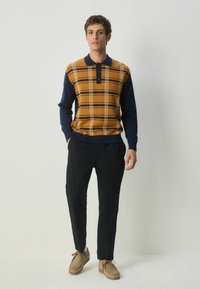 Polo-sweater i sennepsgul med marineblå ærmer, prydet med et ternet mønster, knapper ved kraven, parret med mørke bukser og brune sko.