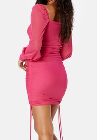 Vestido rosa ajustado con mangas largas de tul, escote cuadrado, detalles fruncidos y lazos ajustables en el dobladillo. Tela suave y elástica.