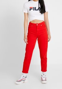 Mujer vistiendo un top corto blanco de FILA, pantalones rojos de cintura alta, zapatillas blancas de FILA, de pie contra un fondo blanco liso.