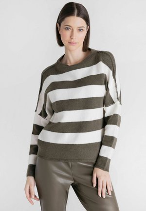 Vrouw met kort donker haar, die een bruin-wit gestreepte trui en bruine leren broek draagt, staand tegen een effen lichte achtergrond.