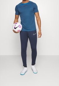 Conjunto deportivo de Nike que incluye una camiseta azul de manga corta con cuello en contraste, combinada con pantalones ajustados azul marino y zapatos de fútbol blancos, sosteniendo un balón de fútbol.