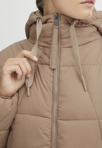 Beige puffer jacket with a high collar and zipper closure. Features a textured fabric, adjustable hood drawstrings, and quilted design.