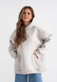 Licht beige oversized hoodie van zacht materiaal, met een grote voorkauw, een capuchon met trekkoord en geribbelde manchetten, gecombineerd met blauwe jeans.