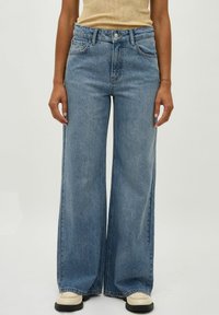 Light blue wide-leg jeans with a high waist, made from denim; features five pockets and subtle fading. Worn with cream shoes.
