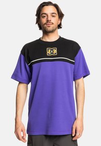 Fialové a černé tričko s kr átkým rukávem, na kterém je žluté logo DC. Bavlněná tkanina s volným střihem a vodorovným detailem bílého pruhu.