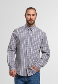 Mann mit Bart und zurückgebundenem Haar, der ein knöpfbares, langärmliges, rot-blau-weiß kariertes Hemd und eine blaue Jeans trägt, mit einer Hand in der Tasche.