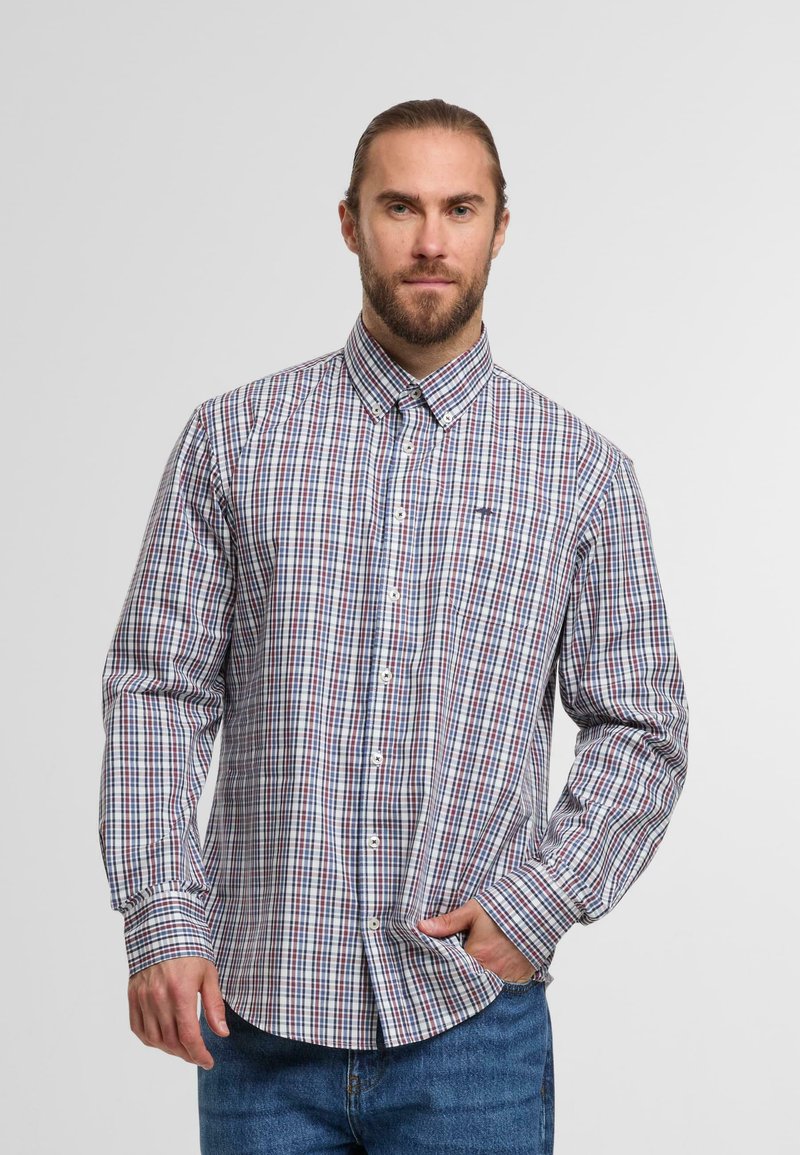 Mann mit Bart und zurückgebundenem Haar, der ein knöpfbares, langärmliges, rot-blau-weiß kariertes Hemd und eine blaue Jeans trägt, mit einer Hand in der Tasche.