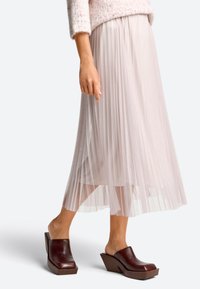 Plissé midi rok in lichtroze stof, gecombineerd met donkerbruine wedge mules. De rok heeft een zachte, vloeiende textuur en een elastische tailleband.
