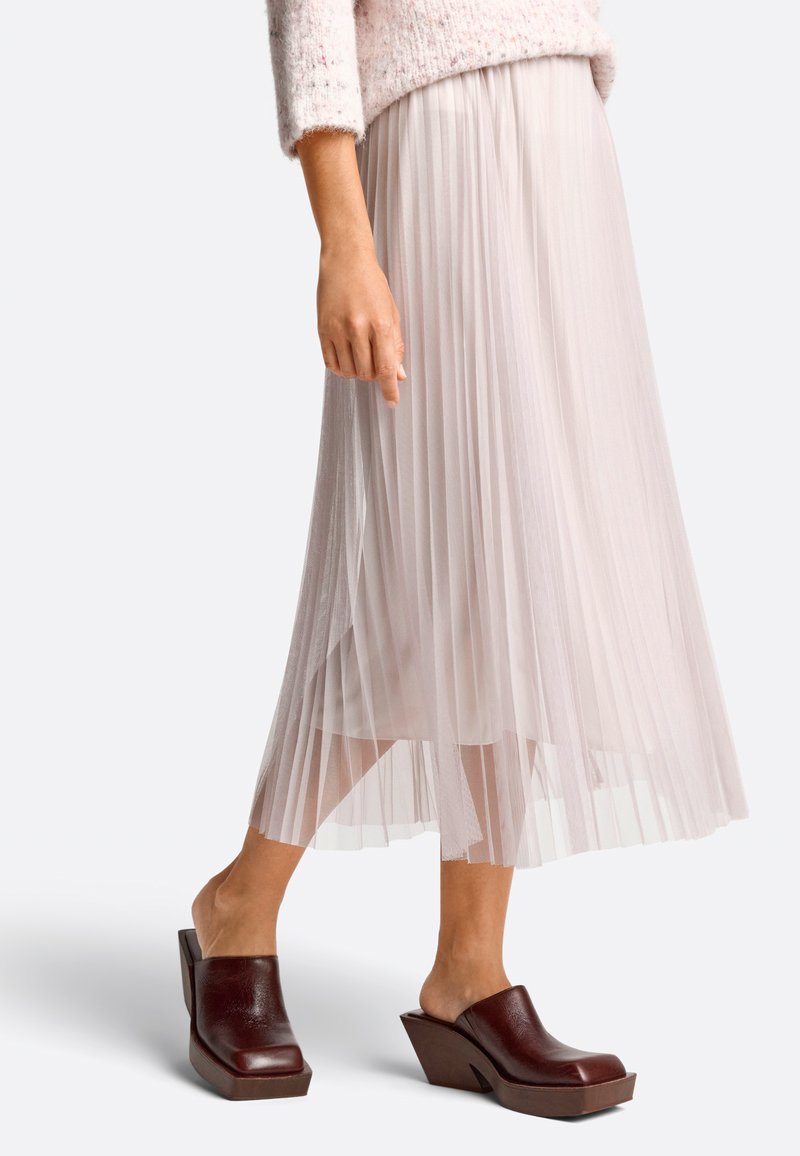 Plissé midi rok in lichtroze stof, gecombineerd met donkerbruine wedge mules. De rok heeft een zachte, vloeiende textuur en een elastische tailleband.
