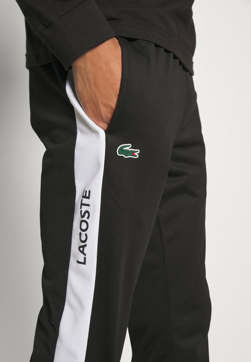 Pantalons de sport noirs avec des bandes latérales blanches, arborant un logo crocodile vert Lacoste. Fabriqués en matériau texturé avec des poches.