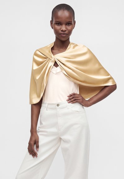 Femme aux cheveux courts portant une cape en satin doré sur un haut blanc et un pantalon blanc taille haute, posant avec une main sur la hanche.