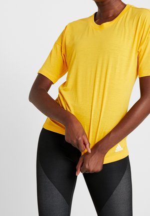 Person iført en gul Adidas t-shirt og sorte teksturerede leggings, der holder i kanten af t-shirten med begge hænder.