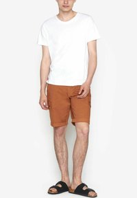 Pantaloni scurți cargo maro cu manșete rulate, buzunare laterale și un tricou alb cu guler rotund. Pantalonii scurți au o textură netedă și o potrivire lejeră.
