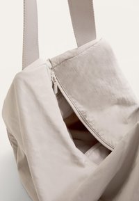 Beige tygväska med dragkedja, med en mjuk, krinklad textur och breda, platta remmar för bekväm bärning.