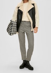 Gilet en simili cuir noir avec doublure en shearling crème, sweat-beige, pantalon pied-de-poule noir et blanc, et bottes chelsea noires à semelle épaisse.