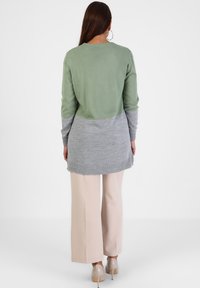 Langer Pullover aus grünem und grauem Strick, mit rundem Ausschnitt, geschwungener Saum und langen Ärmeln, kombiniert mit beigen, weiten Hosen.