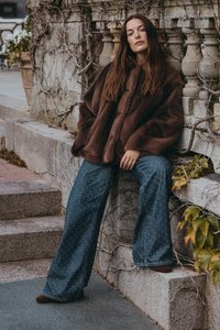 Kvinna med långt brunt hår klädd i en brun pälsjacka och mönstrade byxor med vida ben sitter på stensteg vid ett stenräcke täckt av vinrankor.