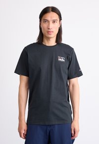 Zwart katoenen T-shirt met korte mouwen, ronde hals en een klein wit logo op de borst. Model staat tegen een effen achtergrond.
