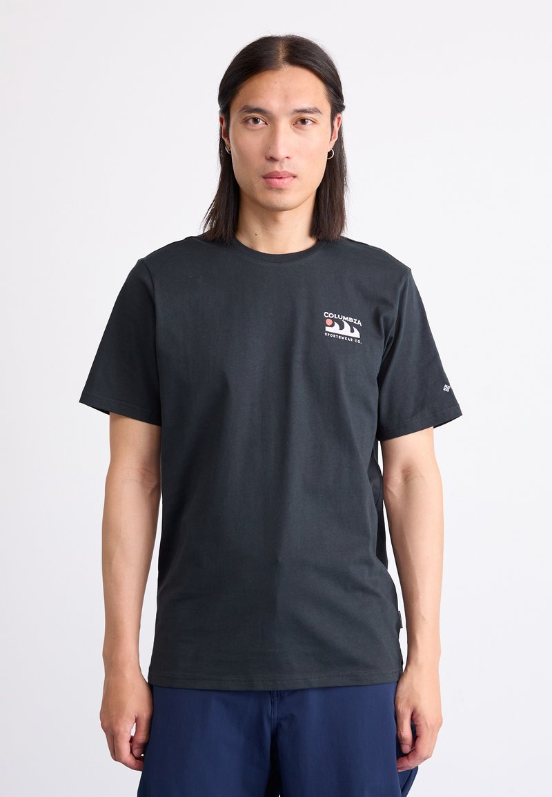 Zwart katoenen T-shirt met korte mouwen, ronde hals en een klein wit logo op de borst. Model staat tegen een effen achtergrond.