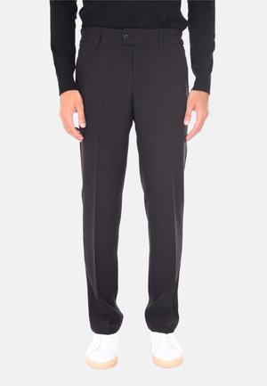 Uomo che indossa pantaloni neri su misura, sneakers bianche e una maglietta nera a maniche lunghe, in piedi contro uno sfondo chiaro.