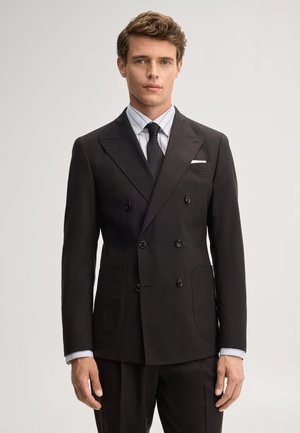 Uomo con abito nero doppiopetto, camicia bianca a righe e cravatta nera, in piedi davanti a uno sfondo chiaro e semplice.