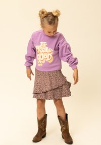 Jong meisje met dubbele knotjes dat een paarse trui draagt met de tekst "It feels good," een bloemenrok en bruine cowboylaarzen, en naar beneden kijkt.