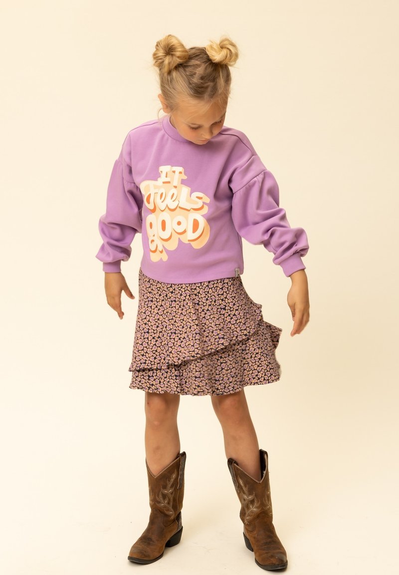 Jong meisje met dubbele knotjes dat een paarse trui draagt met de tekst "It feels good," een bloemenrok en bruine cowboylaarzen, en naar beneden kijkt.