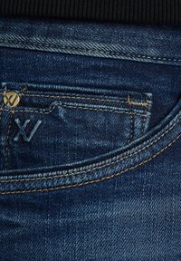 Dunkelblaue Jeans-Taschen mit kontrastierenden goldenen Steppnähten und einem kleinen Metallnieten, die nahe der Taschenkante mit "XJ" geprägt ist.