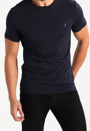 Mand iført en tætsiddende navyblå kortærmet t-shirt med en lille lyseblå logo på venstre bryst, kombineret med sorte bukser.