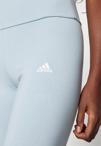 Lichtblauwe sportleggings met ribbeltextuur, voorzien van een wit Adidas-logo op het onderbeen. Close-up van de stof en handdetail.