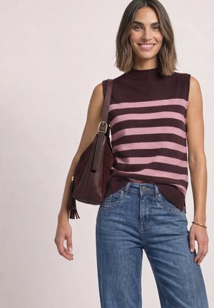 Femme souriante portant un pull sans manches rayé et un jean, portant un sac à bandoulière brun foncé avec détail pompon.