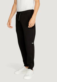Black sweatpants made of soft material with a drawstring waist, tapered cuffs, and side pockets. Features a small logo on the left thigh.