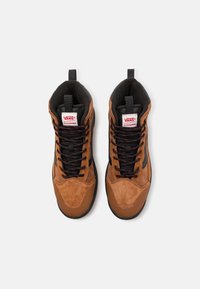 Baskets montantes marron avec des dessus en daim et en textile, cols rembourrés noirs, lacets noirs avec des accents rouges, et un logo Vans sur la languette.