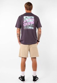 T-shirt en coton gris foncé avec un imprimé graphique multicolore au dos, associé à un short beige, des baskets noires et des chaussettes blanches.