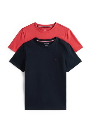 Enfant souriant aux longs cheveux bouclés, debout pieds nus, portant un t-shirt noir et un pantalon rayé gris clair sur fond blanc uni.