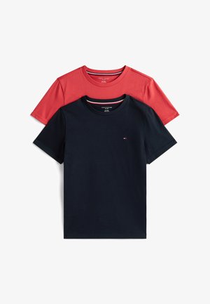 Lachend kind met lang krullend haar staat blootsvoets, draagt een zwart t-shirt en lichtgrijze gestreepte broek tegen een effen witte achtergrond.