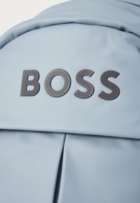 Világos kék hátizsák texturált anyaggal és kiemelkedő sötétszürke "BOSS" logóval. Jellemzője egy sima, lekerekített tetejű és középen varrott rész.