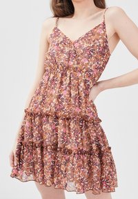 Femme portant une robe sans manches à décolleté en V, avec des motifs floraux dans des tons rose, marron et orange, ornée de volants à niveaux et de fines bretelles.