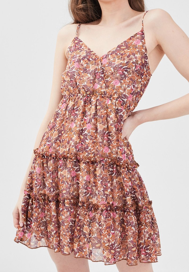 Femme portant une robe sans manches à décolleté en V, avec des motifs floraux dans des tons rose, marron et orange, ornée de volants à niveaux et de fines bretelles.