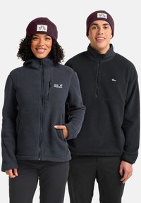 Deux personnes souriantes, portant des vestes en polaire sombres et des bonnets en tricot violets avec des logos Jack Wolfskin, se tenant côte à côte sur un fond blanc.