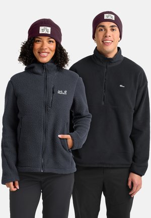 Deux personnes souriantes, portant des vestes en polaire sombres et des bonnets en tricot violets avec des logos Jack Wolfskin, se tenant côte à côte sur un fond blanc.
