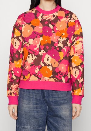 Kvinde iført farverig blomstret sweatshirt i nuancer af pink, orange og brun med ribkanter ved ærmer og bund, kombineret med løse blå cowboybukser.