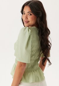 Blusa verde con texture, maniche corte a palloncino, vita arricciata e orlo a peplum; realizzata in un tessuto morbido e leggero con dettagli sottili.