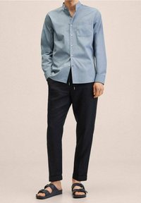 Camicia azzurro chiaro con colletto alla coreana, taschino sul petto, abbinata a pantaloni scuri e sandali neri. Tessuto liscio.